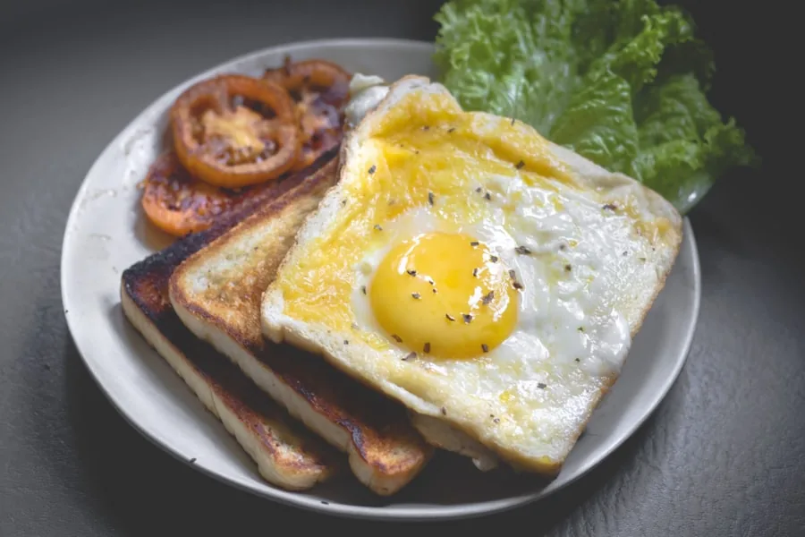 A fried egg cooked into a slice of toast, with two toast triangles, grilled tomato halves and salad leaves on a plate