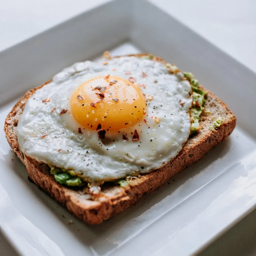 A fried egg with chilli flakes and black pepper on wholegrain avocado toast, on a square white plate