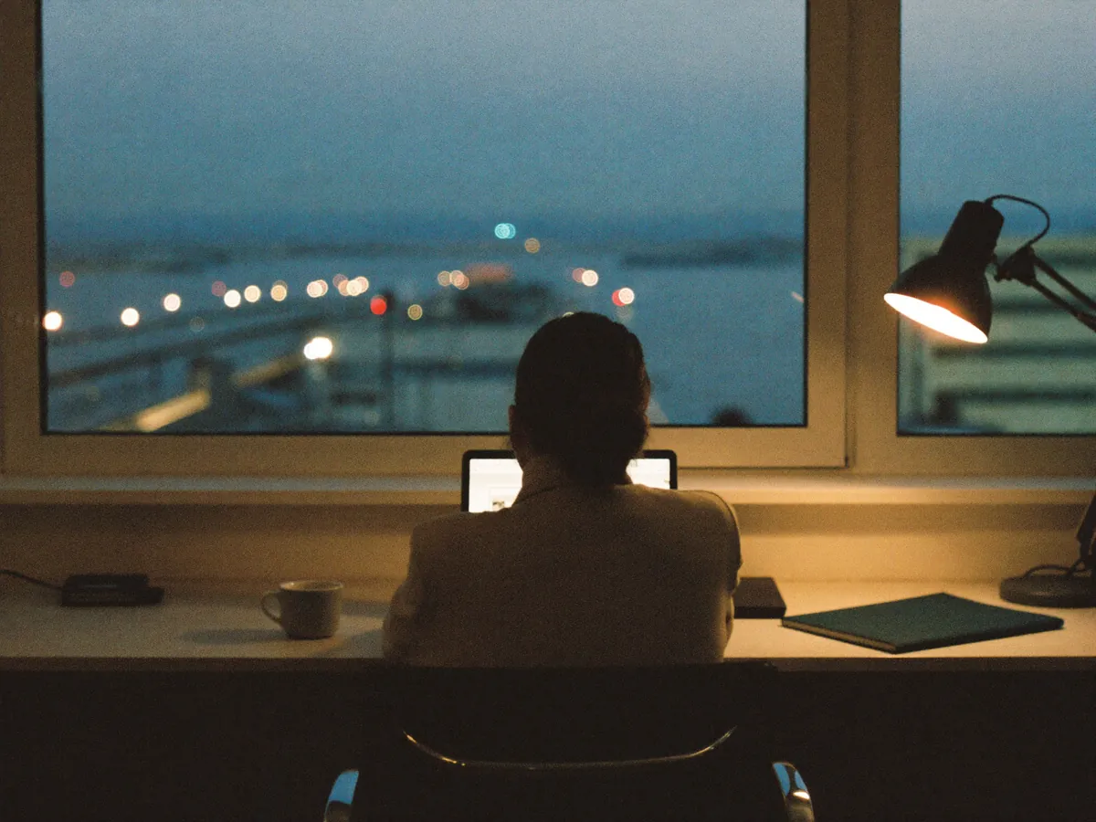 A person at a desk by the window in the evening, with a desk lamp on and city lights outside